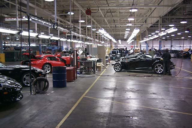 A group of motorcycles parked in front of a building - Pistonheads - The image showcases an industrial setting, likely a car manufacturing or assembly plant. In the foreground, several cars, varying in color and model, are neatly arranged in rows. Some cars are in the process of being worked on, as indicated by a car being cleaned. The space is well-lit with fluorescent lights, possibly coming from an upper level. There are also several people present, all wearing dark clothing, suggesting they may be employees of the factory. The factory's infrastructure, including beams and metal structures, can be seen in the background, contributing to the industrial atmosphere. The ground is made of concrete and is marked by yellow lines, possibly indicating paths for vehicles.