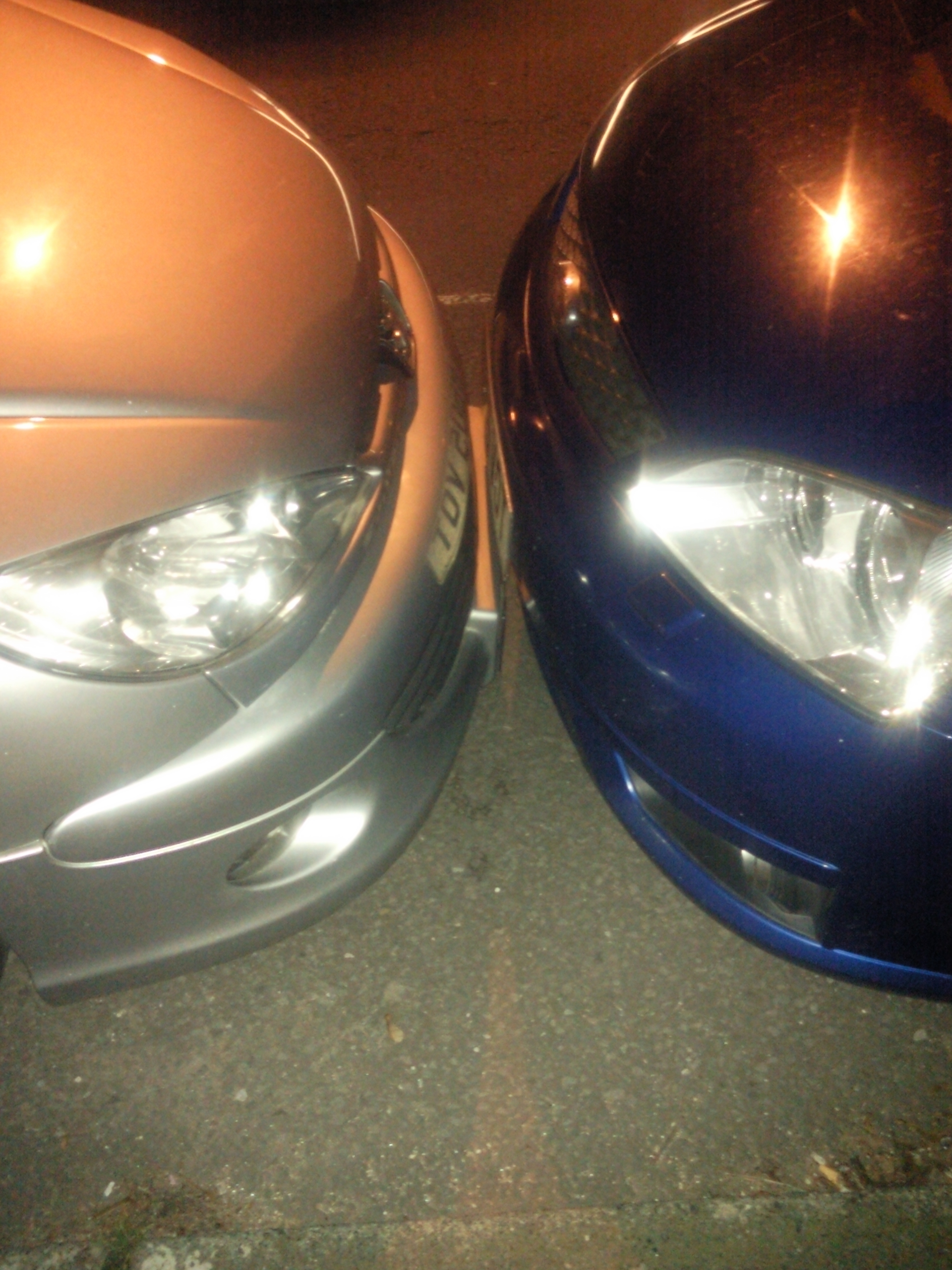 The BAD PARKING thread [vol3] - Page 63 - General Gassing - PistonHeads - The image features two cars parked side by side on a street. The cars appear to be small or compact in size, and they are painted in different colors: one is blue, while the other is orange. The scene is viewed from an angle where the front ends of the vehicles are facing each other. The photo is taken during what appears to be nighttime, suggesting the quietness of the area.