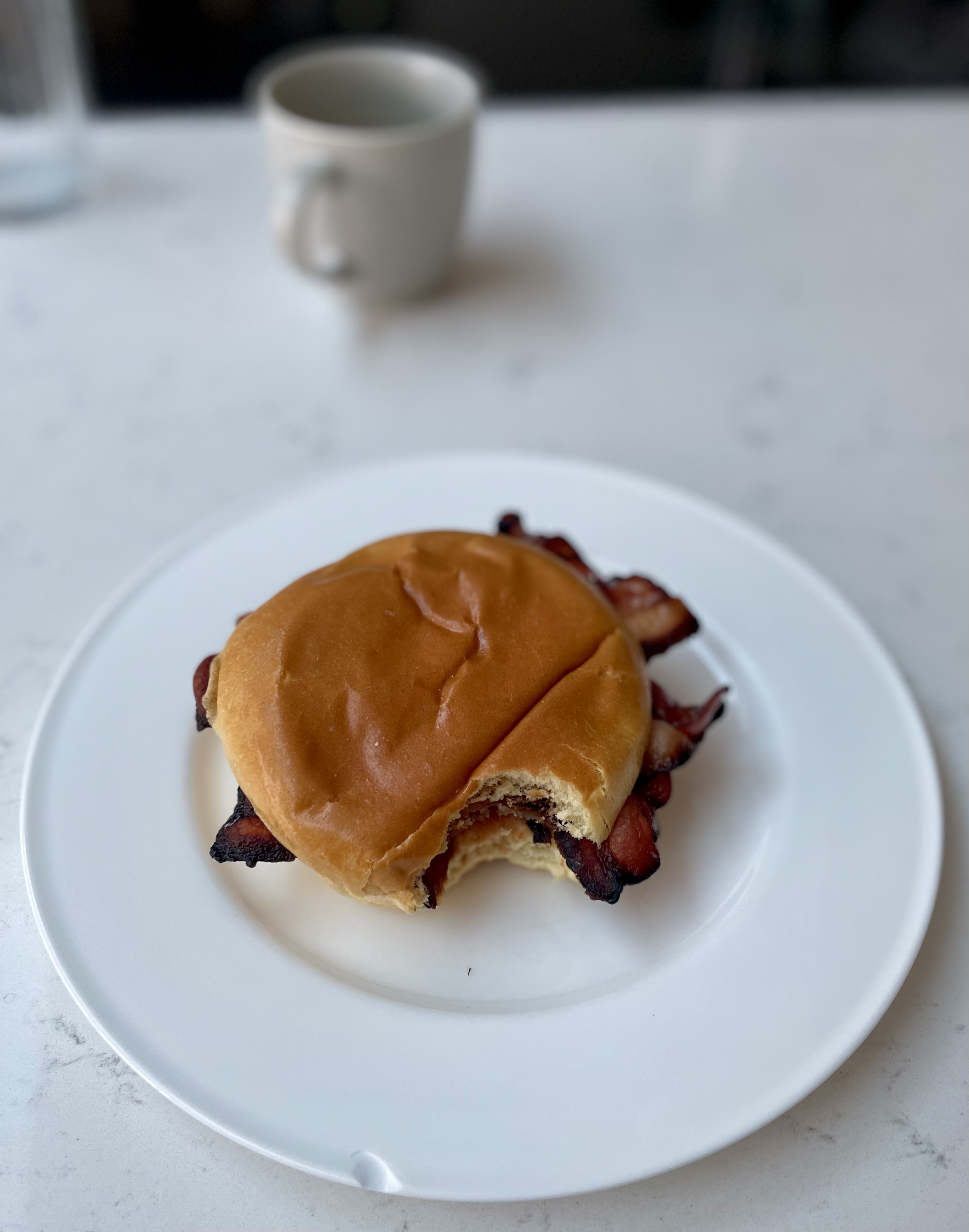 Pistonheads - The image shows a close-up of a breakfast sandwich, which appears to be a bacon sandwich, with a single strip of bacon visible. The sandwich is placed on top of a white plate and is partially wrapped in paper, suggesting it might still be warm. There's a wooden table beneath the plate, and in the background, there are blurred objects that suggest a casual dining environment. On the table to the left, there's a coffee cup with a handle, indicating that the meal is accompanied by a hot beverage. The overall ambiance of the image suggests a relaxed and simple morning routine.