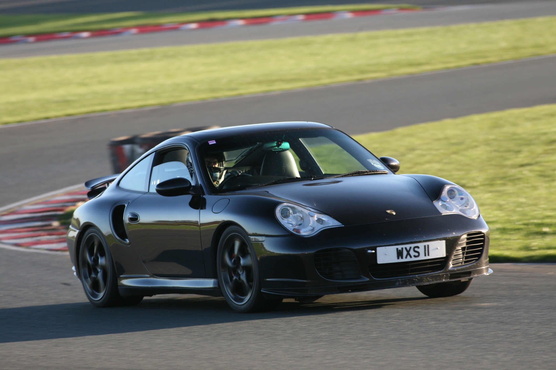 Shall we have a PH Top 100 - Best 4wd performance cars - Page 6 - General Gassing - PistonHeads - The image shows a dark colored sports car driving on what appears to be a race track. The car is seen at an angle, facing slightly upwards, indicating motion. There are no visible people or signs in the photo, and the focus is solely on the car and its immediate environment. The car is moving along a section of the track that also curves gently around a grassy area. The race track has a black top with red and white markers along the edges. The image has a dynamic feel due to the car's speed and the angle of the shot.