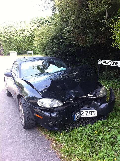 A car is parked on the side of the road - Pistonheads - The image shows a dark-colored sports car with significant damage to its front. The car has sustained large hits, causing the hood to be almost entirely ripped off. The front grille and bumper are also damaged or missing. The car is parked in what appears to be a natural setting with lush green grass and dense, leafy trees. On the right side of the image, there is a sign posted that has the word "Downtow" on it. The overall scene suggests an accident or impact that took place, particularly given the state of the car and the context of the natural environment and sign.