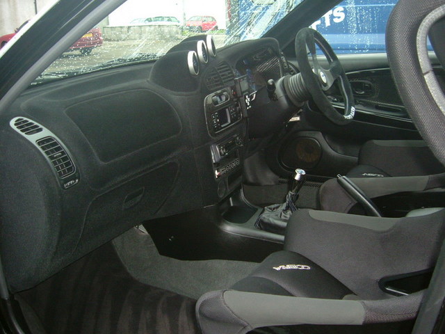 Registeredpics Evo Newley Pistonheads - This is a black and white photograph taken from the perspective of a vehicle's interior. The foreground shows the driver's side of the car, featuring the steering wheel, gear shift, and various controls within the center console. Beyond the seats, the interior of the car is visible, and through the windshield, the storage compartment of a truck can be seen outside. The image captures the sleek and modern design of the car's motorsport-inspired interior.