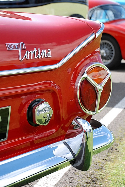 Goodwood Breakfast Club, Pre 1966 classic sunday - Page 1 - Goodwood Events - PistonHeads - The image captures a close-up view of the back of a vibrant red car. The car features a white frame license plate, indicating that it might be of European origin. Dominating the scene is a bright red gemstone-shaped emblem with the word "Cortina" written on it. The overall design suggests a classic or vintage style car, possibly a model from the 1960s or earlier. The image exudes a sense of nostalgia and automotive history.