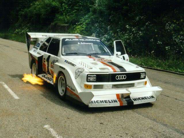 Pistonheads - In the image, there is a white Audi race car in full motion on a rural road. The car has a prominently displayed yellow logo on its side. The road is lined by trees, and there is a grassy area visible to the left of the car. The car is generating an impressive amount of heat by oiling on the tarmac, as evidenced by the flames being visibly emitted from the rear.