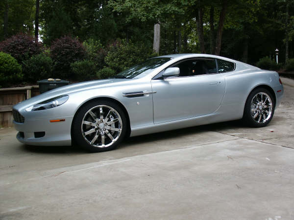 The Proper Colour for an Aston thread - silver, greys, black - Page 1 - Aston Martin - PistonHeads - The image shows a silver sports car parked in a concrete driveway. It features distinctive alloy wheels, a streamlined body, and a visible convertible top. The car is well-maintained with a polished appearance. The surrounding area appears to be a residential setting with green foliage and a natural backdrop, giving the impression of a leisurely, tree-lined street.