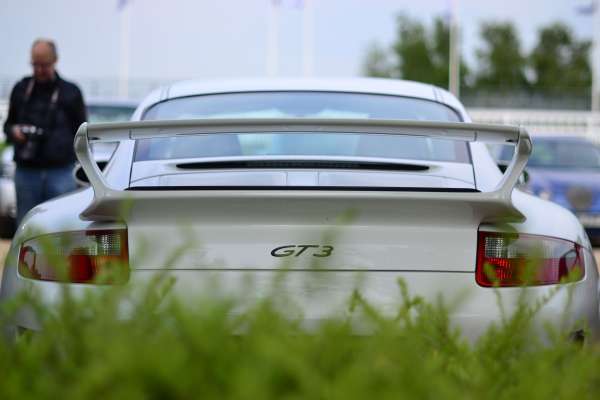 In addition to the cars, there's a man standing who is partially visible from the waist up and is wearing a black jacket. The rear view of the car in focus reveals it to be a Porsche GT3, identifiable by the text and logo on the tail. The car is parked on a grass field, and the background shows other cars and a few trees. The lighting could suggest either dawn or dusk, and the sky is partly cloudy, enhancing the overall mood of the scene.