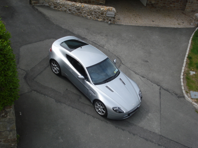 No regrets - Page 1 - Aston Martin - PistonHeads - The image showcases a silver sports car parked on a paved, curved street. The vehicle's sleek design and shiny finish reflect the sunlight, suggesting a clear day. The background consists of a grassy area, a building, and a stone wall, providing a contrast to the modernity of the car. The smooth lines of the car and its shiny surface suggest a well-maintained vehicle, while the surrounding scenery suggests a residential area.