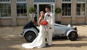 Soon be a 7 owner and just saying hi. - Page 1 - Caterham - PistonHeads - The image depicts a couple posing for a picture on a brick-paved surface. The woman, dressed in a white bridal gown, holds a bouquet of red flowers, while the man is attired in a traditional white formal outfit with a boutonniere. They are standing next to an Aston Martin, which is parked near a large windowed building with white frames. The scene suggests a formal event, likely a wedding, as implied by the attire and the red bouquet. The Aston Martin, though not the focus, adds a touch of elegance to the photograph.