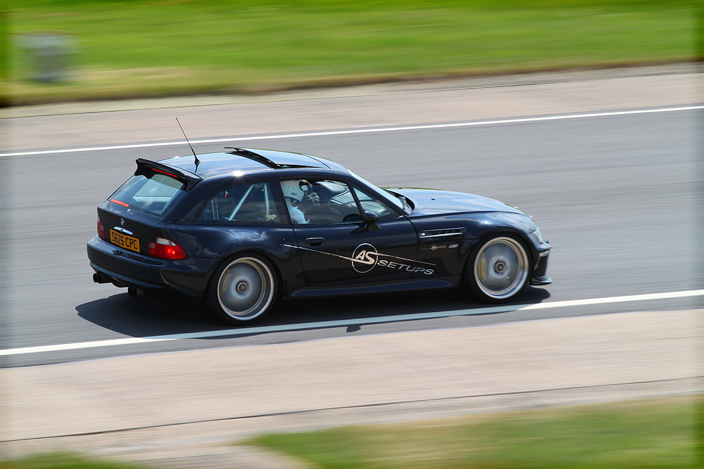 Your Best Trackday Action Photo Please - Page 5 - Track Days - PistonHeads - The image is a dynamic photograph of a two-door black sports car captured in motion on a divided highway. The vehicle displays a spoiler and a rear wing, along with a prominent badge, suggesting it might be a BMW, given its design and styling. There are blurs of greenery to the right of the car, indicating the speed of the vehicle as it moves along the road. The wheels and the road surface exhibit a radial pattern, indicating a fast turn. The photograph captures the action and the motion of the car with a sense of speed and motion.