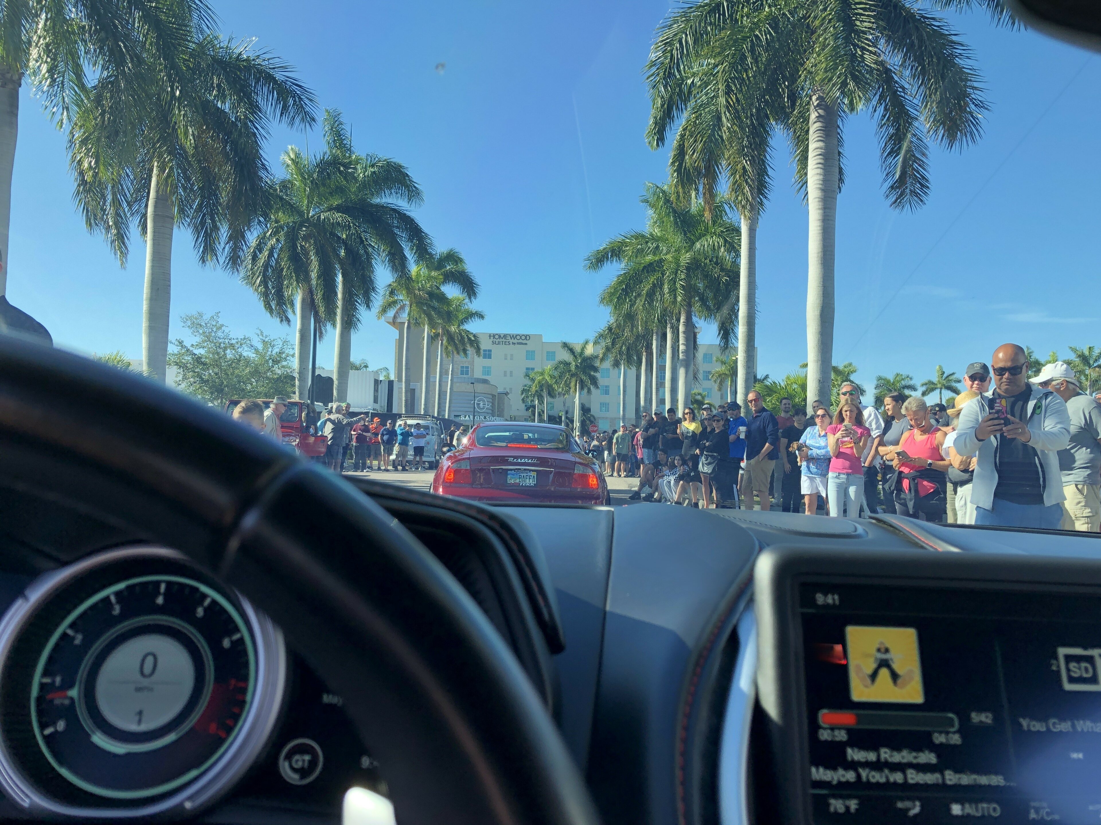 Pistonheads - The image shows a city street scene during daylight. In the foreground, there is a close-up of a vehicle's interior, with a steering wheel visible and the dashboard showing an array of indicators and gauges. Beyond this, the view extends to the outside world where numerous people are walking along a sidewalk, likely in a tourist area given the presence of palm trees. The street is lined with parked cars, and the sky is clear blue, indicating fair weather. In the background, there's an urban setting with buildings and a clear sky. There's no text visible on the image.