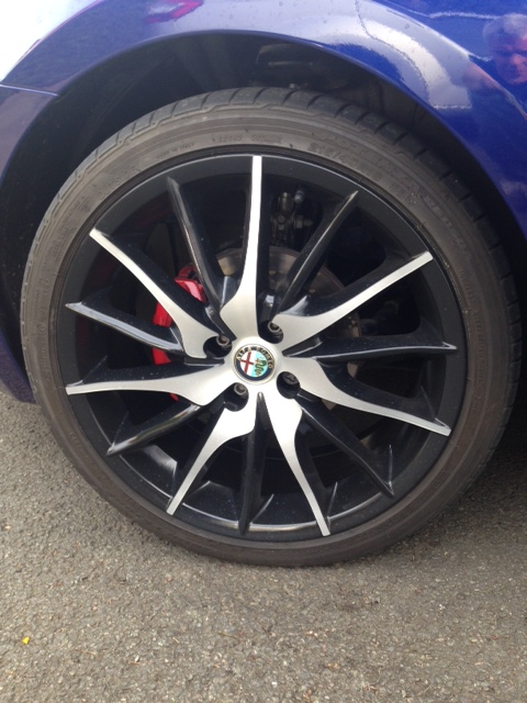 Alloy Wheel Restore and Minor Paint Repair in Northants? - Page 1 - Northamptonshire - PistonHeads - The image is a close-up photograph focusing on the wheel of a car. At the center, there is a distinctive emblem, resembling a shield, surrounded by the spokes of the wheel. The wheel itself has a deep color with the rims and spokes appearing to be shiny, contrasting with the matte color of the wheel's surface. The inside of the wheel is marred by a visible scratch on the lower right side. The car is parked on a surface that appears to be an asphalt road.