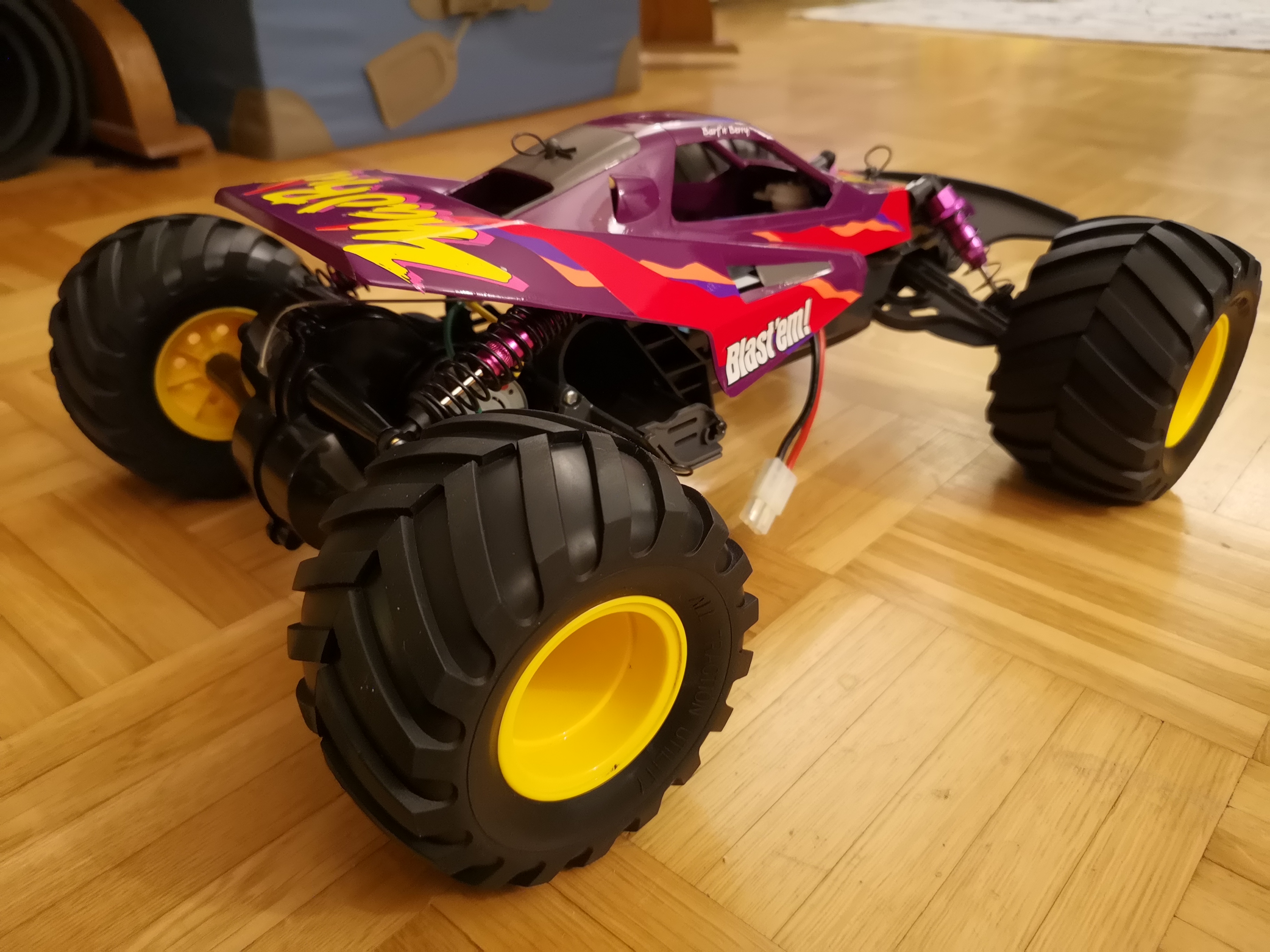 Tamiya for [-]my son[-] me - Page 1 - Scale Models - PistonHeads - The image shows a vibrant toy monster truck on a wooden floor. The truck is predominantly purple with accents of yellow and red, and it features large wheels typical of monster trucks. The cab of the truck is painted in a contrasting color, possibly blue or purple, adding to its eye-catching design. On the side of the truck, there's visible text, but it's not clear enough to read. There's a single figure in the background, which appears to be a human standing and facing away from the camera. The style of the image is a straightforward photograph with no additional text or graphics on it.