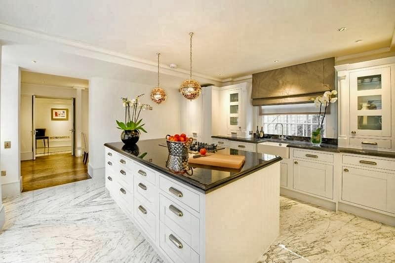 Which Kitchen Worktop? Pros and cons? - Page 6 - Homes, Gardens and DIY - PistonHeads - This image captures a spacious and well-decorated kitchen. The kitchen is large with ample counter space and white cabinetry. A glass-fronted cabinet on the right side of the image displays a pristine white interior. On the kitchen island, there is a bowl filled with what appears to be red fruit, surrounded by various potted plants. Above the island, a modern chandelier hangs, and a vase with flowers adds a touch of color and life to the space. The floor is covered in tiles, adding to the clean and luxurious aesthetic of the kitchen.