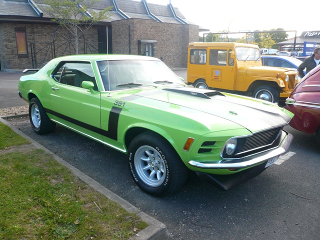 Pistonheads Metrocentre - The image features a striking green and black Mustang muscle car parked in a lot alongside other regular cars. The Mustang, parked on the left side of the image, has a chrome grill, side mirrors, and dual exhaust. The Mustang's hood is adorned with a black stripe and graphics. In the background, there is a building, possibly a gas station or restaurant, with another vintage vehicle parked beside it. The sky is overcast, suggesting that the photo was taken on a cloudy day.