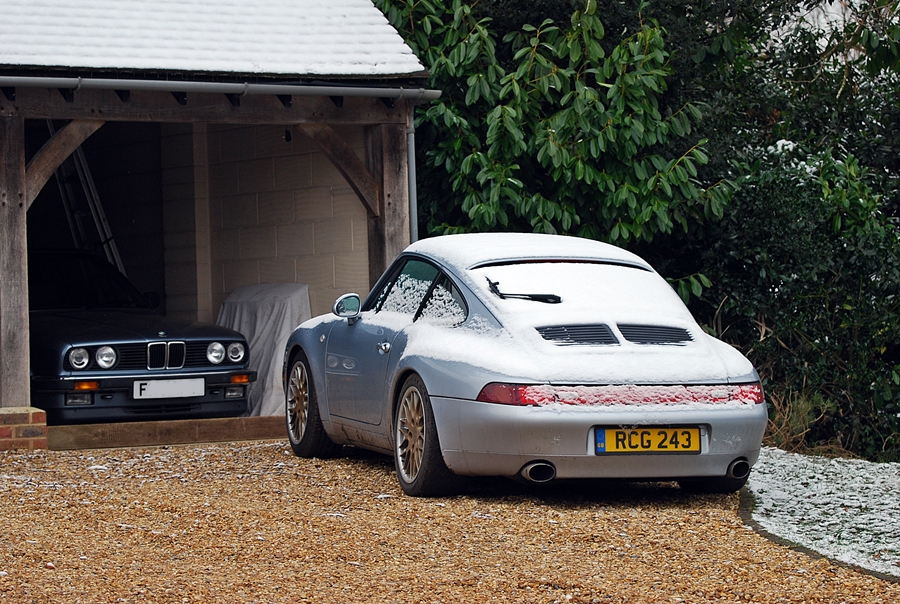 A picture a day... Porsche - Page 32 - Porsche General - PistonHeads - The image features a tranquil outdoor scene with two cars parked in front of a barn or shed. On the left, there's an older model car that appears to be a convertible, while on the right, there's a more modern sports car with its hood open, revealing a snowboard secured under it. The sky is overcast, suggesting either recent rainfall or an impending change in weather. The vehicles are parked on a gravel driveway, and both cars have snowflake designs on their windows. In the background, there's a glimpse of a person who seems to be working near the barn. There are no texts visible in the image.