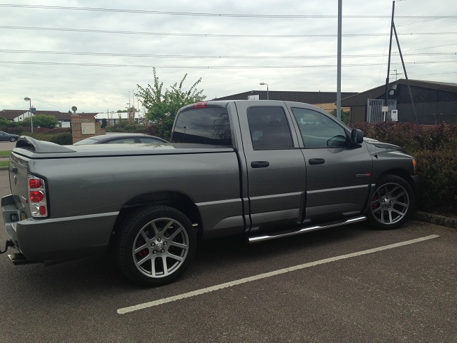 Wanted....Dodge Ram SRT 10 - Page 3 - Yank Motors - PistonHeads - The image depicts a gray Dodge Ram truck parked in a parking lot. The truck is viewed from the rear, showing its tailgate, rear lights, and windows. It appears to be a double cab model with a full bed and a towing package. The consistent color and without any visible customization, it looks like a standard model from a recent year. The truck is on pavement, with line markings indicating parking spaces. In the background, there are power lines and some overgrown shrubbery, suggesting an outdoor or semi-urban setting.