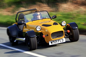 A red motorcycle is parked in a parking lot - Pistonheads - The image shows a person driving a small, three-wheeled car. The car is predominantly yellow with black accents, and it has a distinctive face design on the front. The vehicle is in motion on a road, as indicated by the blurred background suggesting speed. The license plate on the car reads "W32FLD," and the person driving appears to be wearing sunglasses and a helmet for safety. The setting appears to be a rural or suburban area during daylight, given the natural light and lack of urban structures.