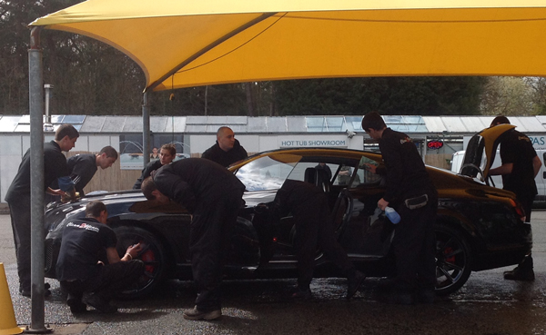 Clean in 60 Seconds... - Page 1 - Rolls Royce & Bentley - PistonHeads - The image shows a group of men in black outfits engrossed in work on a black sports car parked under a yellow tent. The men are huddled together, leaning over the car, possibly attending to maintenance tasks. Some of them are using tools, and a couple of bottles, likely containing fluids or care products, are visible near them. In the background, there's a sign that reads "HOT TUB SHOWERoom," suggesting the setting might be an event or a service station with facilities for car care and repair. There are also other cars and trucks parked in the vicinity. Overall, the image captures a moment of teamwork and dedication in what appears to be a professional car care situation.