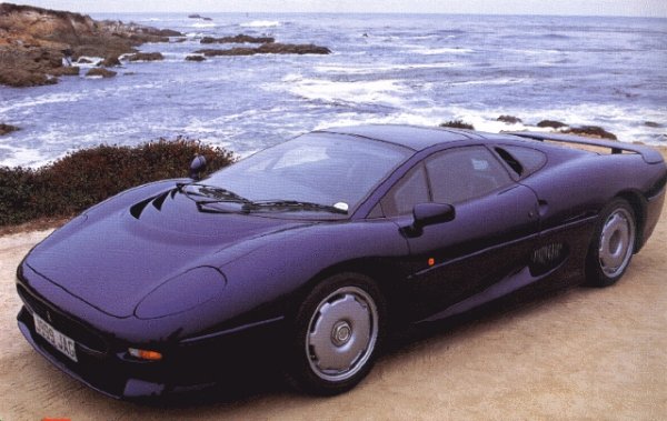 Pistonheads Year Birth - The image features a convertible sports car with a black finish, sitting on a plain, sandy surface. The car is positioned front-right, showcasing its front end and part of the driver-side door. The background is dominated by an expansive view of the ocean, with waves splashing against the rocks along the shoreline. The sky is overcast, providing a moody atmosphere. The overall setting suggests a coastal area, possibly a deserted beach given the absence of other vehicles or people.