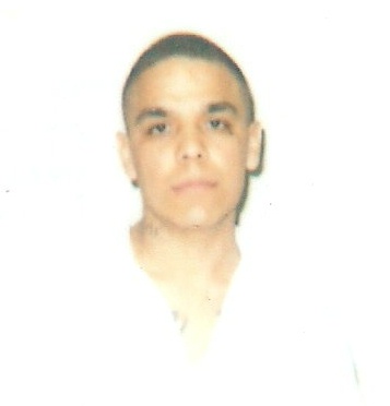 The image appears to be a photographic portrait of a person. The individual has short hair, and is wearing what seems to be a white collared shirt. The background is plain and lacks distinguishing features. The person has a neutral expression and is looking directly at the camera. The lighting on the person's face is even, and the image does not provide any additional context or setting information.