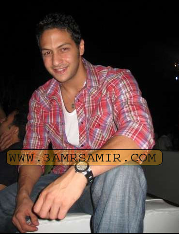 The image is a photograph of a smiling man sitting on what appears to be a white wall or surface. He is wearing a casual, short-sleeved shirt with a mix of plaid patterns and also has a watch on his left wrist. The lighting suggests the photo was taken in the evening or night, as the background is dark. No people are visible in the immediate background, keeping the focus on the individual in the foreground. Since the image also contains text, such as 'WWW', '3AMRSAMIR.COM', it can be inferred that this image is likely for commercial purposes, possibly related to a website.