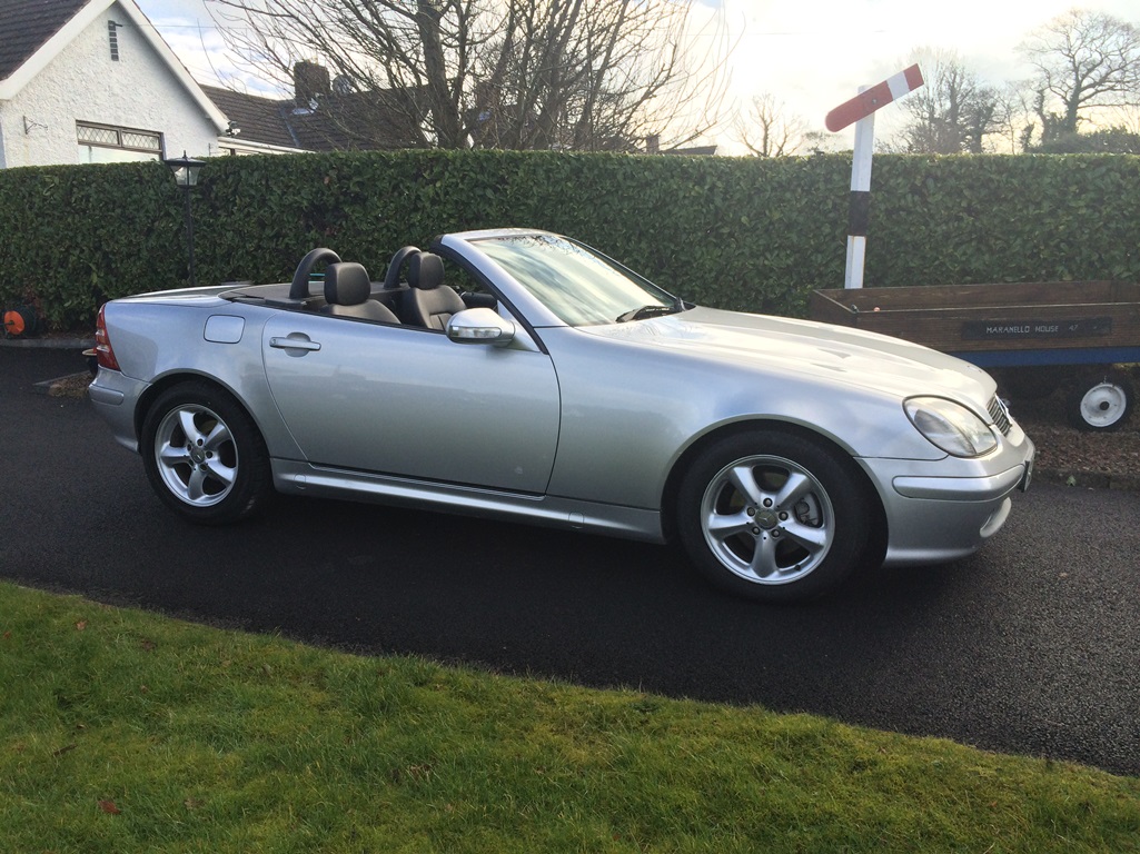 Lets see a picture of your classic(s) - Page 248 - Classic Cars and Yesterday's Heroes - PistonHeads - The image showcases a silver Mercedes C-Class convertible car parked in a driveway in front of a house. The car is in motion with the top down, and there are three visible seats. The speedometer indicates the vehicle is traveling at 12 miles per hour. The car is positioned in front of a blue wooden trailer and a red and white traffic sign indicating "do not enter" to the left of the image.