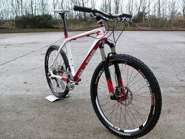 The "Show off your bike" thread! - Page 352 - Pedal Powered - PistonHeads - The image showcases a close-up of a mountain bike standing on a hard surface. The bike has a predominantly white and red color scheme, with the frame's color transitioning towards black at the bottom. The front disc brake and the rear derailleur are both clearly visible. A kickstand is present, providing support to the bike. The backdrop of trees indicates an outdoor setting, possibly a park or recreational area.