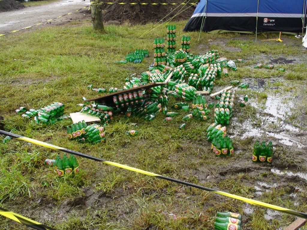 Beer Mountain in style  - Page 1 - Le Mans - PistonHeads - The image depicts a large quantity of glass bottles, most of which appear to be beer bottles, strewn across the grass in a concentrated pile. Many of the bottles are empty and broken, suggesting a recent abandonment. In the background, there is a tent pitched on grass, possibly indicating a festival or outdoor event. There is a black and white rope strung across the grass, with cones at each end, indicating a boundary or safety zone.