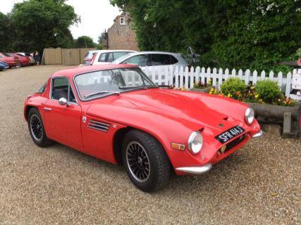 Early TVR Pictures - Page 55 - Classics - PistonHeads - The image showcases a vibrant red vintage car, which is parked on a gravel driveway. The car's design suggests it's from the 1950s or 1960s, featuring iconic details associated with classic sports cars. The gravel driveway is bordered by a white picket fence, adding a charming, suburban feel to the scene. The background reveals the outlines of parked cars and a wooden house, suggesting a residential setting. The image communicates a sense of nostalgia through the contrasting elements of the vintage car and the modern, suburban environment.