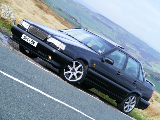 Pistonheads - The image features a black sports car parked on the side of a road, overlooking a scenic landscape. The car's sleek design and large alloy wheels suggest it is a high-performance model. The license plate reads "NI4 LBM". The background includes a lush green hill, a rocky outcropping, and a quaint village nestled in a valley below the car's vantage point. The crisscrossing of lights and shadows on the car's hood indicates it might be a sunny day. There are no people visible in the image.