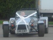 Soon be a 7 owner and just saying hi. - Page 1 - Caterham - PistonHeads - The image features a vintage car, prominently displayed in the foreground. It has a distinctive silver color and is adorned with a white ribbon running across its window frame. The car is equipped with black fenders and round, orange-rimmed lamps. The car is parked on a roadway, and in the background, a portion of a delivery truck is visible, partially obscured by the car. The overall scene suggests a special event or perhaps a street parade, given the decoration on the car's window.