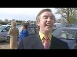 A man and a woman standing next to each other - Pistonheads - The image is a still shot from a video that appears to be a scene from a reality TV show or documentary film. It shows a Caucasian man in the foreground wearing a suit with a yellow shirt and a decorated red tie. His expression is one of surprise or amusement, with his mouth open and his eyes focused to the side. In the background, there are two more individuals who are less clear but seem to be in conversation; one is wearing a red top and the other has gray hair. They are standing outdoors, near vehicles parked in an orderly manner, suggesting this scene might be taking place in a parking lot or a similar public area. The environment looks overcast with gray skies, indicating it might be a cloudy day.