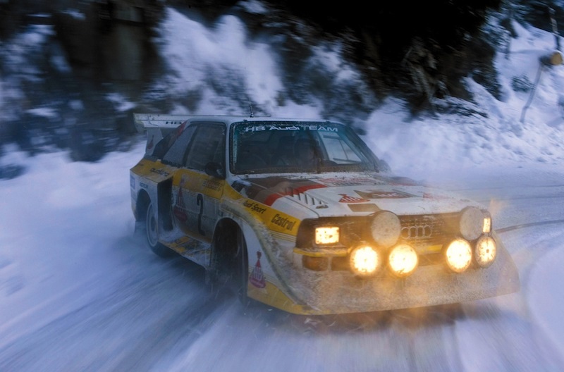 Just awesome vehicle-related images. - Page 4 - General Gassing - PistonHeads - The image shows a snowy mountain road where a car is driving, kicking up a cloud of snow. The car appears to be a rally or race car, given its small, compact size and the number "99" visible on its side. The vehicles headlights are on, illuminating the path ahead. In the background, the landscape is covered in snow, and we can see the dense vegetation typical of mountainous terrain.