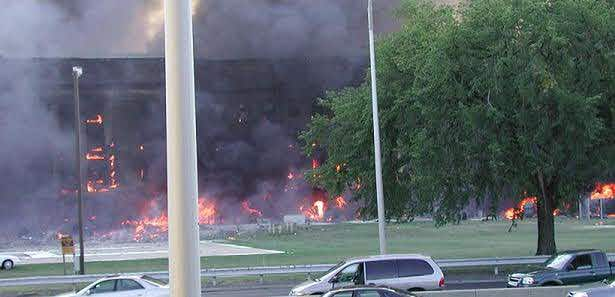 The image captures a scene of destruction with a building engulfed in flames. Thick black smoke billows from the burning structure, indicating intense heat and fire. The building is surrounded by trees, adding a touch of greenery to the otherwise devastating scene. On the road in front of the building, several cars are neatly parked, oblivious to the chaos unfolding behind them. The level of detail and the contrast of the vehicles against the burning building paint a stark picture of the situation.