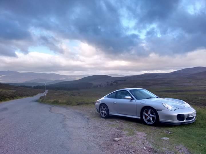 The road to a 911 996 C4S (Alfa Romeo, MG & GTI Content)  - Page 1 - Readers' Cars - PistonHeads - The image showcases a silver Porsche coupe parked on the side of a winding road with a backdrop of green mountains under a cloudy sky. The car is positioned on what appears to be the edge of a green hillside, overlooking a network of roads. The vehicle's sleek design contrasts with the natural surroundings. The color palette is dominated by shades of green, gray, and blue, creating a serene and picturesque scene.