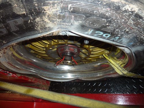 Aftermarket rims for the Radical? - Page 1 - Radical - PistonHeads - The image shows a close-up view of the wheel well of a vehicle. Focus is on the hubcap and tire, and the tire appears to be a Goodyear with a visible tread pattern. There is a yellow cable attached to the wheel, and the wheel is set inside a red vehicle frame. The overall condition of the tire and wheel suggests alloy or steel construction with some dirt or dust covering the face of the tire. The cable is used for lifting or lowering the tire, which is indicated by the presence of a hoist to assist in the correct positioning.