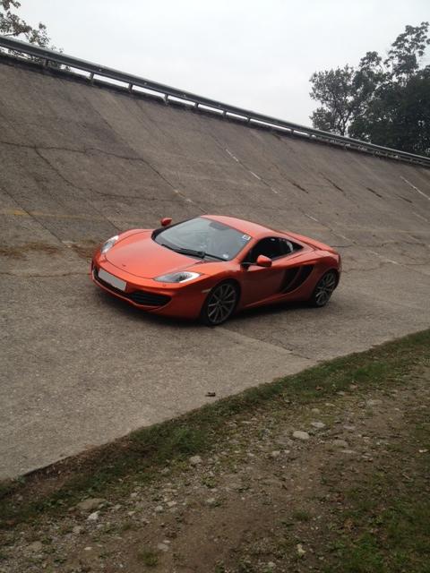 Welcome... - Page 1 - McLaren - PistonHeads - The image shows a bright orange Bentley GT2 RS car parked on a steep concrete surface, which appears to be part of a race track given the presence of what looks like a runoff area and a gravel-covered embankment to the right. The car's sleek design and blue and orange tissue box on the dashboard are visible. The background reveals a cloudy sky and trees, suggesting an outdoor setting.