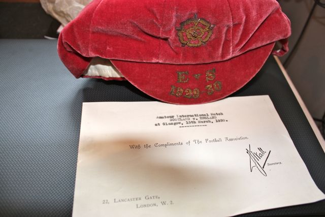 Have these got any value/history? - Page 1 - Sports - PistonHeads - The image shows a red hat with a crest on it, placed atop a document on a black surface. The hat has text on it, reading "E. & S. 1829-30," indicating it could be a hat from that era. Beneath the hat, there's a paper with a shadow cast, detailing a document titled "With the Complements of the Lancaster Gate Football Association." The document commemorates an event that occurred on the 25th of March in the year 1833. The paper has text at the bottom that reads, "S.D. McSpy, Secretary." The hat and document are arranged closely together, suggesting they were placed there intentionally. The black surface underneath provides a stark contrast to the colorful items, making them stand out.