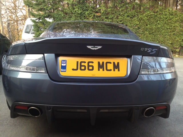 DBS wheels on DB9 - (more or less) - Page 1 - Aston Martin - PistonHeads - The image depicts the rear view of a gray Aston Martin car. The car has a license plate that reads "J66 MCM." The vehicle is seen from behind, with the car parked, isolated, and highlighted by its rear window. The Aston Martin emblem and the registration marker suggest that the car is a luxury and possibly a sports vehicle. The overall setting and the static presentation give a sense of the car's design and identity.