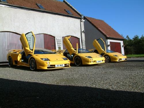 Pistonheads - The image depicts a scene with three yellow Lotus sports cars parked outside a large garage or building. The cars are open, with the hoods popped up, revealing their engines. The setting is casual, with gravel covering the ground and a bright sky overhead suggesting it is daytime. There is a sense of luxury and performance associated with the cars, which are likely sports cars, given their design and demeanor.