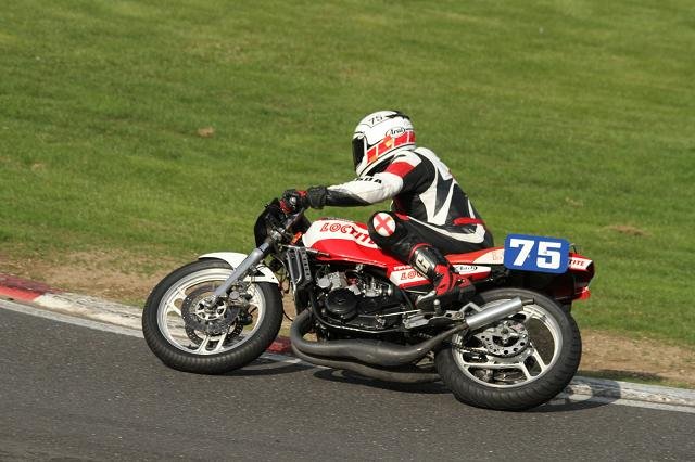 Racers of the '80's - Page 1 - Biker Banter - PistonHeads - The image depicts a motorcyclist on a closed racing track. He is in the process of making a turn with his red and white motorcycle, practicing at high speeds. The rider is wearing full protective gear, including a helmet with visor down, a red and white suit, gloves, and boots. The number 75 is clearly visible on the motorcycle, suggesting it might be used for racing. The background shows the well-maintained surface and a clear sky, indicating a day of good weather.