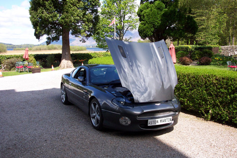 Free Photoshoot Introduction Pistonheads - The image showcases a black car with its hood wide open, parked on a gravel driveway. The environment is lush with trees and hedges surrounding the area, extending to the background where a body of water or lake can be spotted. The partially sunny day adds a pleasant ambiance to the scene. In the far distance, a mountain can be glimpsed against the sky, providing a picturesque backdrop to the opened hood of the car.