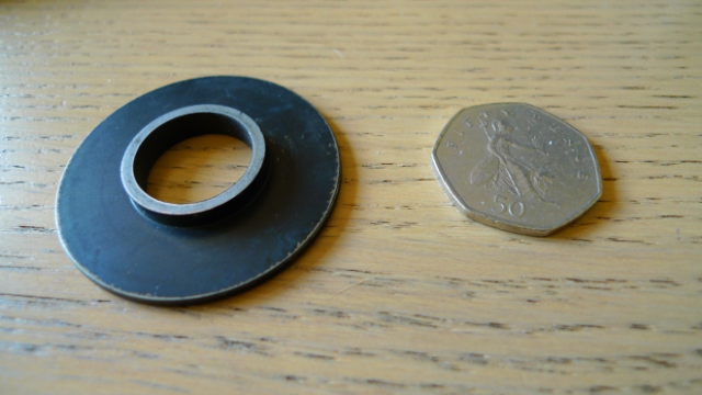 Elise Pistonheads Part - This image features a 50p coin and a small black rubber ring. The 50p coin is placed on a wooden surface, offering a clear view of its mint and embossed symbols. The rubber ring, with its circular shape and a hole in the middle, resembles a stopper or a grommet. The two objects are small in size and are set against a simple, nondescript background. The image presents a stark contrast between the metallic sheen of the coin and the smooth, black texture of the rubber ring.