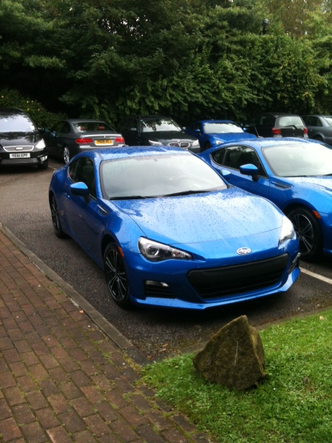 Pistonheads - The image features a striking blue sports car parked in a lot filled with other cars. The blue car is glossy, with a distinct grille and angular design, suggesting it's a newer model. The vehicle is parked adjacent to a small boulder. The lot appears to be a public parking area, as various other cars are visible in the background. The corner of a green patch of grass is visible in the foreground of the image.