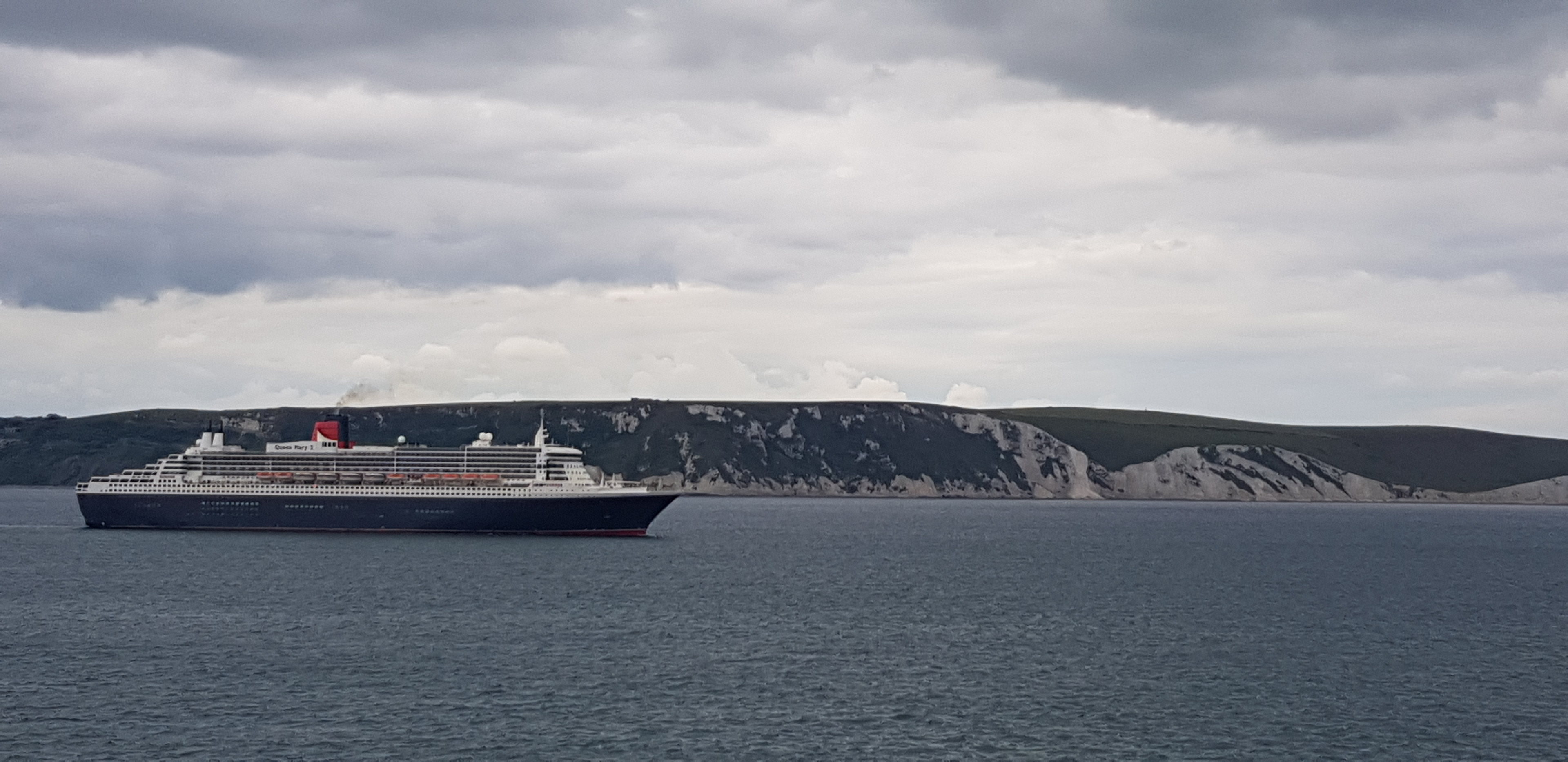 Cruise ships in Weymouth Bay - Page 4 - Boats, Planes & Trains - PistonHeads - The image showcases a large cruise ship sailing on the ocean with a picturesque backdrop of mountains. The sky overhead is cloudy, casting an overcast light over the scene. In the foreground, there's a smaller landmass visible, likely an island or part of the coastline. The contrast between the expansive water and the solid landmass adds depth to the image. The ship is in the center of the frame, its size and position making it a prominent feature of the composition.