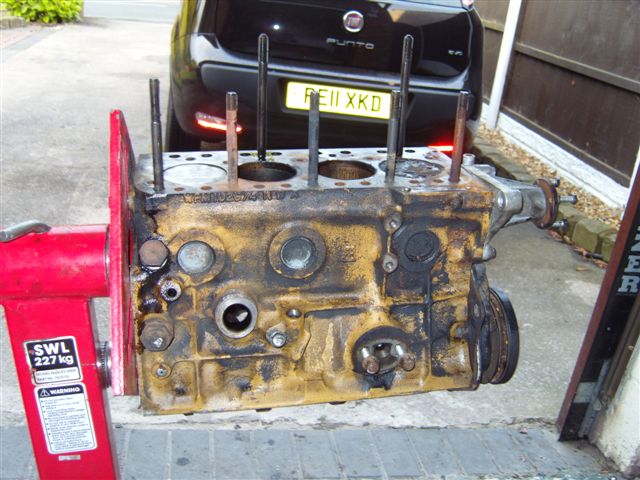 pics - Page 1 - Classic Minis - PistonHeads - The image features a rusted engine block sitting outdoors on a sidewalk, in front of a metal rack with a spare tire mounted on it. The engine has several protruding parts, steeped in black oil and possibly tools used to modify it. There is also a license plate visible at the top of the photo. Nearby, part of a car is visible, suggesting that the viewer might be seeing a snapshot of a garage or a repair process in progress. The image has a sense of stillness, with no people or significant movement captured.