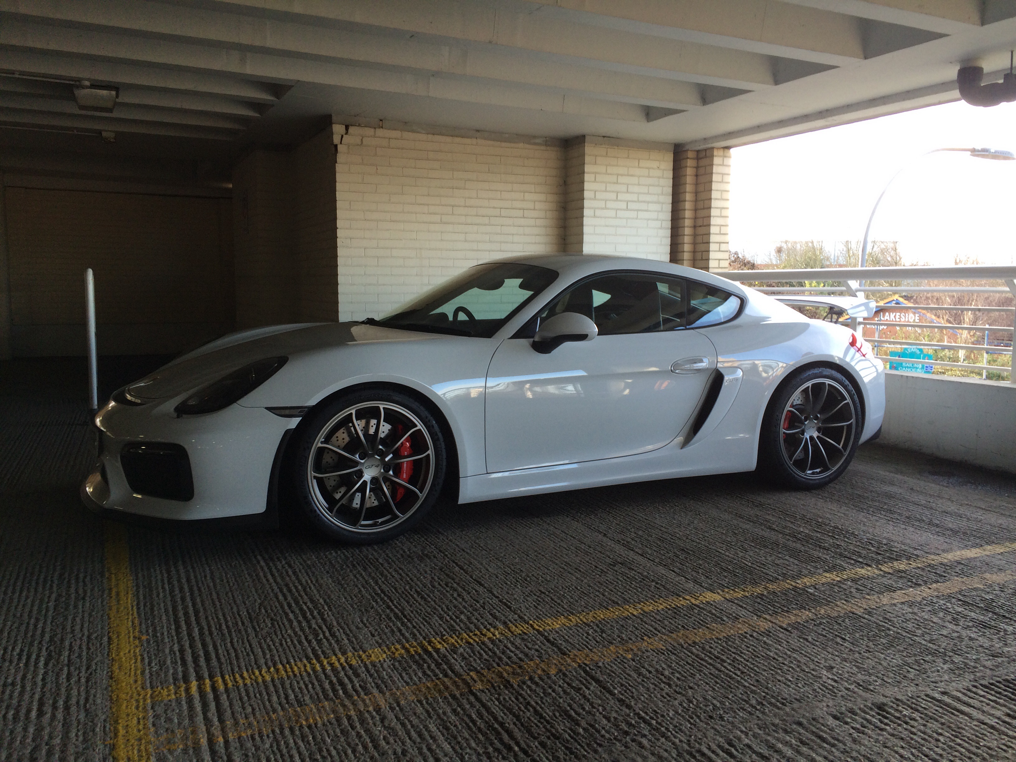 Cayman GT4 delivery and photos thread - Page 36 - Porsche General - PistonHeads - The image depicts a white Porsche sports car prominently parked on a concrete surface within a covered parking structure. The car is positioned at a 45-degree angle to the camera, showcasing its sleek design and alloy wheels. The setting appears to be a mostly empty parking lot, possibly in a suburban area, with a metal railing visible to the right and a bright light source illuminating the scene in the background.