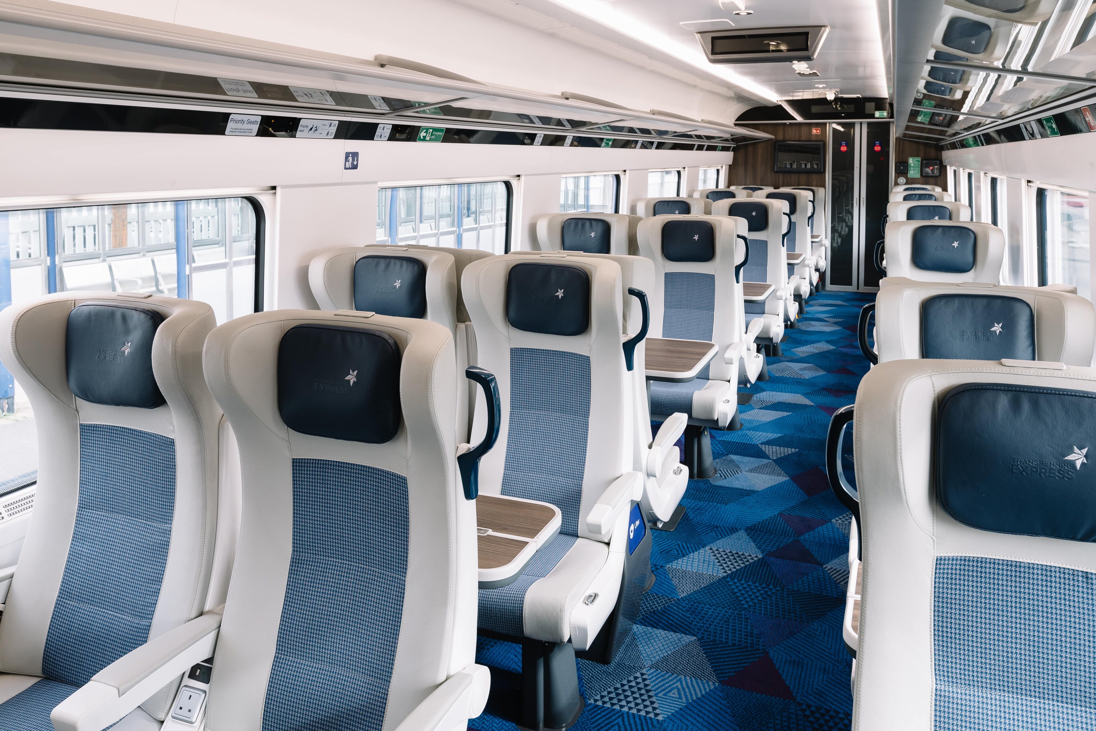 Train coaches overcrowded - Page 2 - Boats, Planes & Trains - PistonHeads UK - The image captures an interior scene of a modern train car. The car is spacious and features blue and white upholstery on the seats, which are arranged in rows facing each other. Overhead, a blue ceiling is adorned with white circular lights, providing ample illumination throughout the space. A large window on one side lets in natural light, enhancing the brightness of the car. The overall ambiance of the train car suggests a comfortable and inviting environment for passengers during their journey.