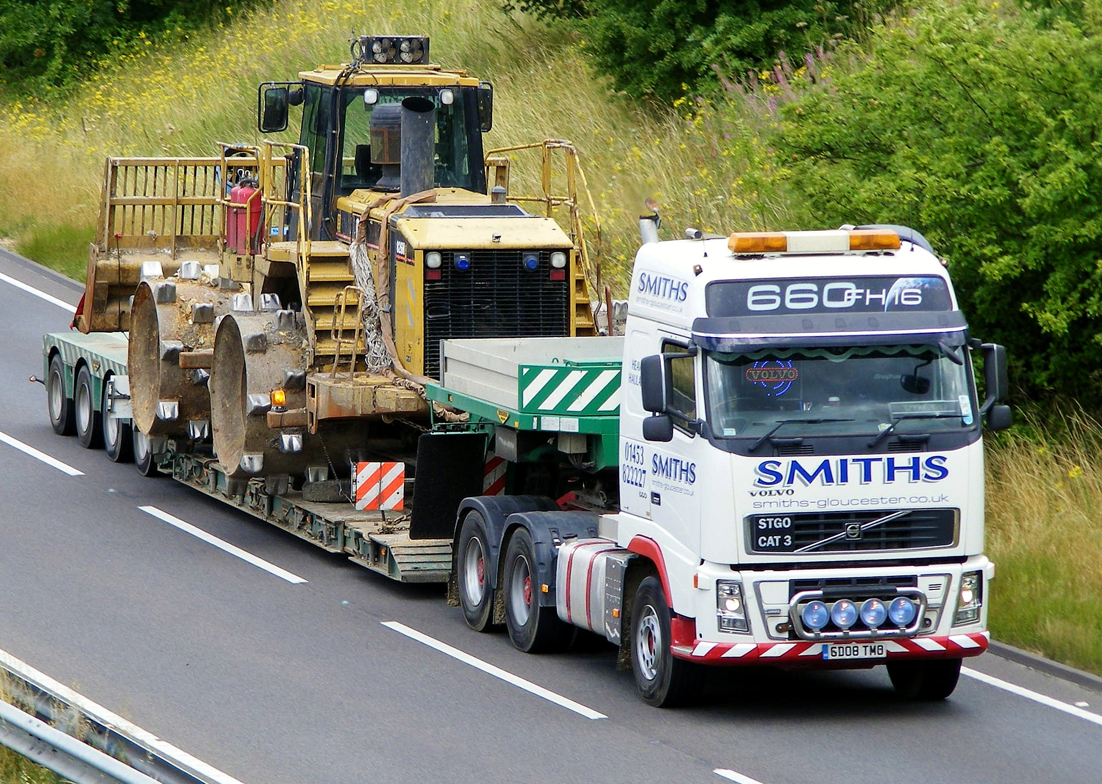 The Heavy Haulage Appreciation Thread - Page 8 - Commercial Break - PistonHeads - The image portrays a scene of industrial activity on a road amidst a natural setting. A large, yellow Caterpillar tractor-trailer rig is towing a green and gray trailer loaded with construction materials. The truck, marked with the number "666" and a phone number, is driving on the right side of the road, with a forest behind it. Various pieces of heavy machinery can be seen in the trailer, suggesting that this could be a temporary transport solution for a construction project.