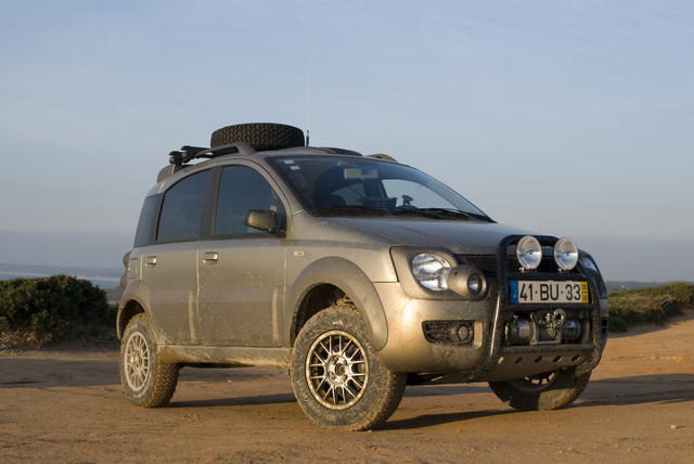 Pistonheads - The image shows a dark-colored SUV on an open dirt surface with a clear sky in the background. The vehicle is equipped with bicycle racks on the roof and two spare tires mounted there as well. It features a prominent, high-clearance bumper with additional off-road accessories and has clear headlights and tail lights. The license plate is attached to the bumper and displays characters and numbers that are not fully visible.